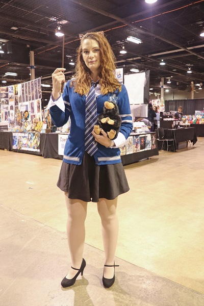 Holding a stuffed Niffler toy and a wand, this cosplayer wears a blue Ravenclaw sweater and poses in front of a vendor.