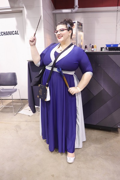 In front of a vendor, this cosplayer is wearing a long blue dress with silver accents running down the sides and along the neckline.