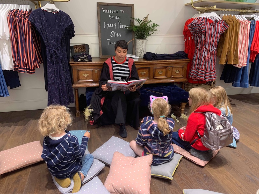 A women dressed in Harry Potter robes reads a chapter of "Harry Potter and the Philosopher's Stone" to a group of children who are sitting on the floor.