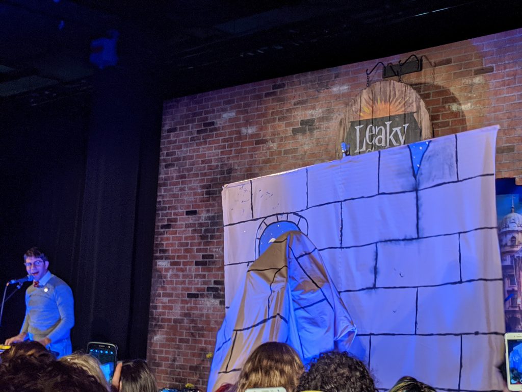 Harry is standing in front of a prop wall and is under a piece of fabric from the same prop wall, making him "invisible."