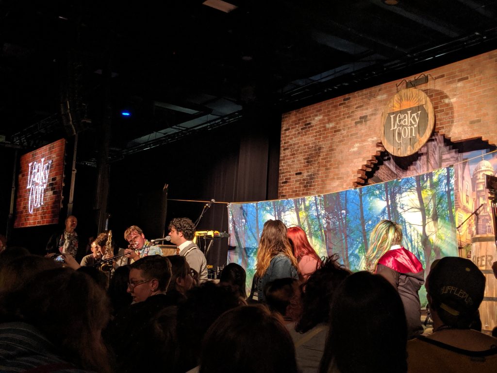 The band is sitting at the edge of the stage with a forest background behind them. The audience is also sitting.