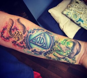A person's arm sports an elaborate and colorful tattoo featuring a snitch, deathly hallows symbol, and stylized dark mark