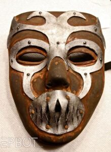 A mask painted rusty brown with lighter shading around the silver swirling pattern laid over the top. Details have been added to make the silver swirling overlay look like metal bolted to the mask.