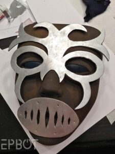 A full-face mask painted with shades of dark brown. Over top of the mask is laid a piece of silver foam cut into a swirling pattern with cutouts around the eyes and a darker-colered grate over the mouth.