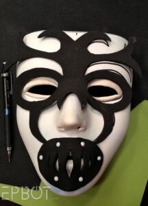 A plain white full-face mask. Over it is laid a piece of black foam cut into a swirling symmetrical pattern with cutouts around the eyes and a grate over the mouth.