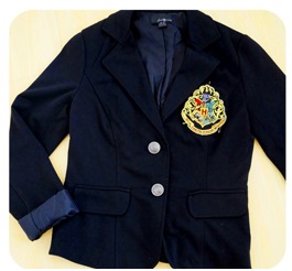 A plain black blazer laid flat and photographed from above. A large Hogwarts school crest badge is positioned on the breast of the blazer.