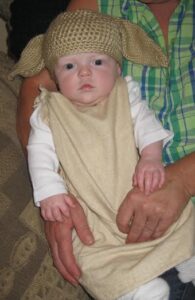 a baby dressed with Dobby the House-elf, in a brown pillowcase-like garment and a homemade hat crocheted to look like elf ears