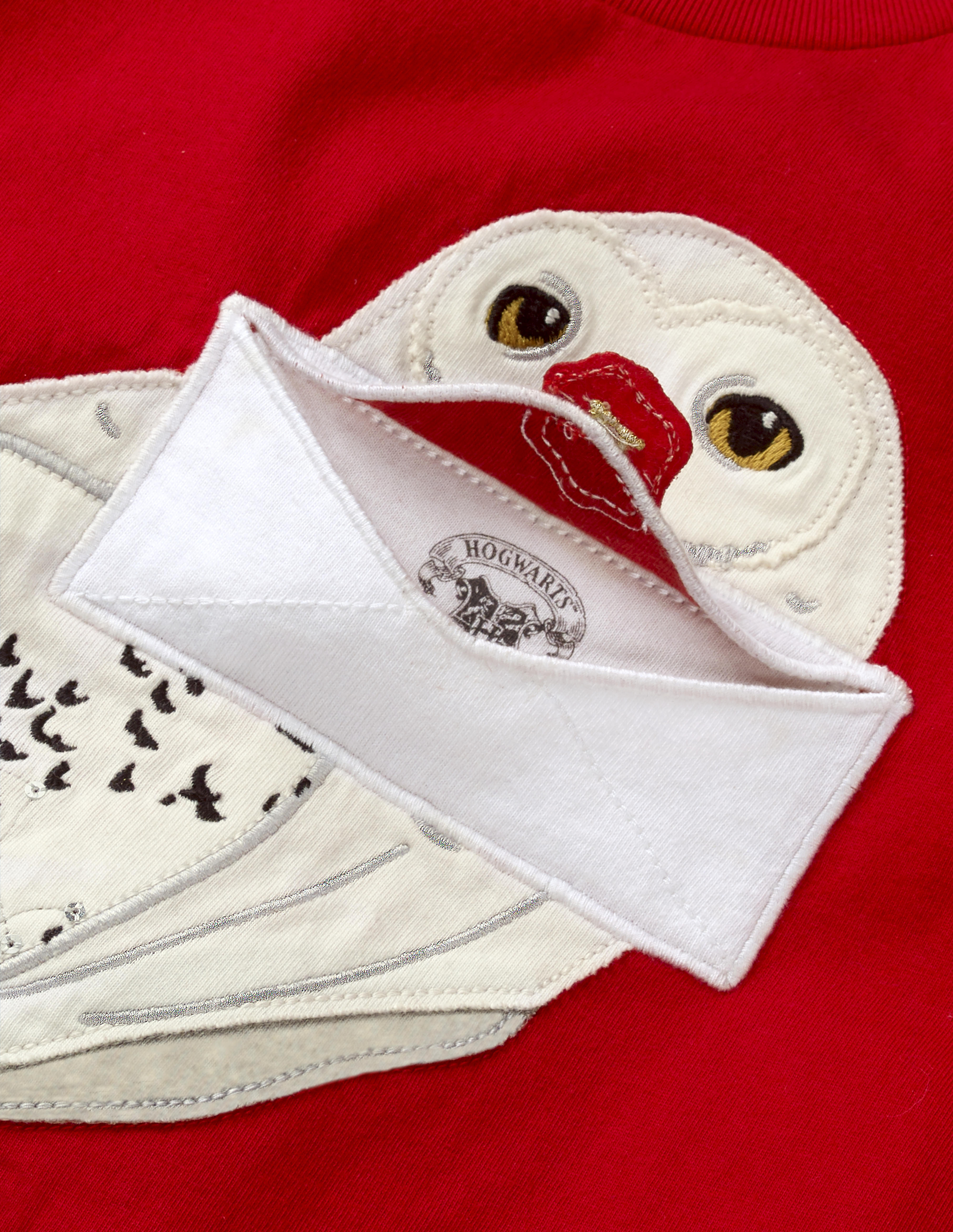 This close-up of the Mini Boden Hedwig Appliqué T-shirt in red shows how the flap of the envelope containing the Hogwarts acceptance letter lifts up to reveal the top of the Hogwarts seal. The shirt retails at £22.