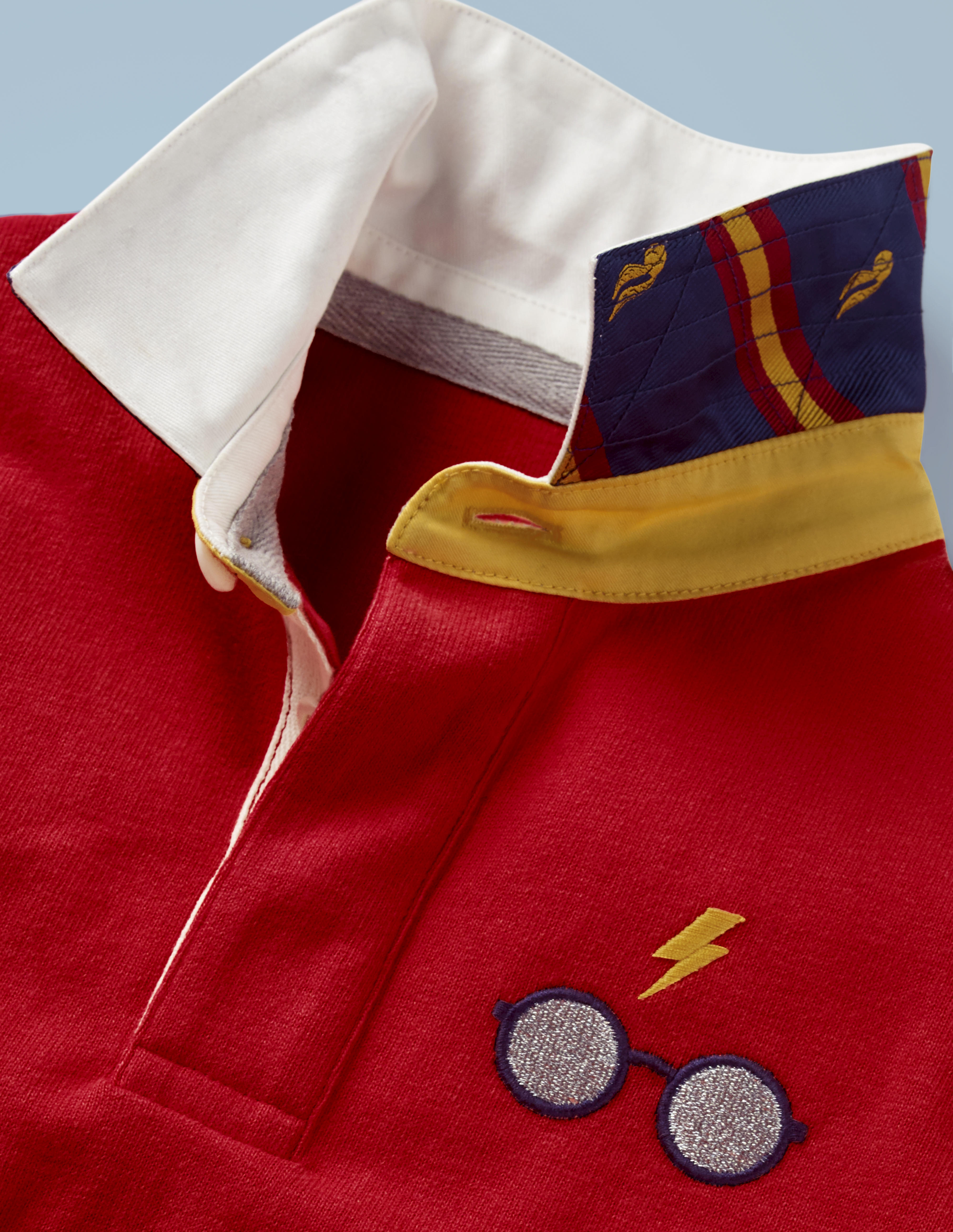 This close-up of the Mini Boden Hogwarts Rugby Shirt in red provides a better look at the collar design and glasses-and-scar logo in the front pocket area. It retails at £30.