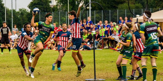 Player in green jersey is holding quaffle and he's trying to score. There is one person in front of him and in font of hoops. There is another two players in green jersey and three players in red-blue jerseys.