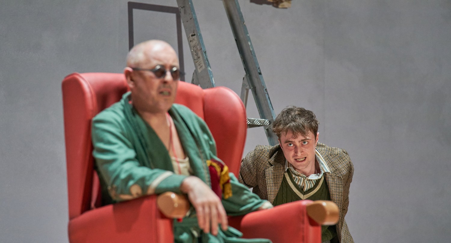 Daniel Radcliffe and Alan Cumming in Endgame on stage. Alan is in a chair as blind and disheveled Hamm while Daniel is looking at him with a mad expression.