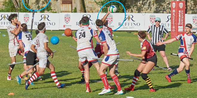 Match UK team vs. team in red-white jersey. From UK team there are male keeper, female chaser, male chaser and male beater. From red-white team there are male keeper, male chaser in the middle with quaffle, male beater with bludger, male chaser and one another player. There are also two hoops in backgroung (belongs to red-white team propably) and one referee in background.