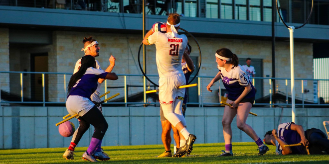 One male chaser in white jersey trying to score quaffle, one other male chaser as support on left, two chasers (one female) from opposite team in blue jerseys trying to block him, one female beater in blue jersey on left, one male beater lying on the ground on right