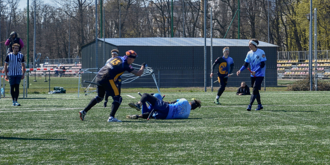 Beater Cerebro from Vienna Vanguard is beating chaser in blue jerseys who is lying on the ground. Propably another player in blue jersey is lying next to him. There is one chaser in blue jersey walking to them. On the backgroung there is one chaser who is running, one referee and someone behind camera for live-stream.