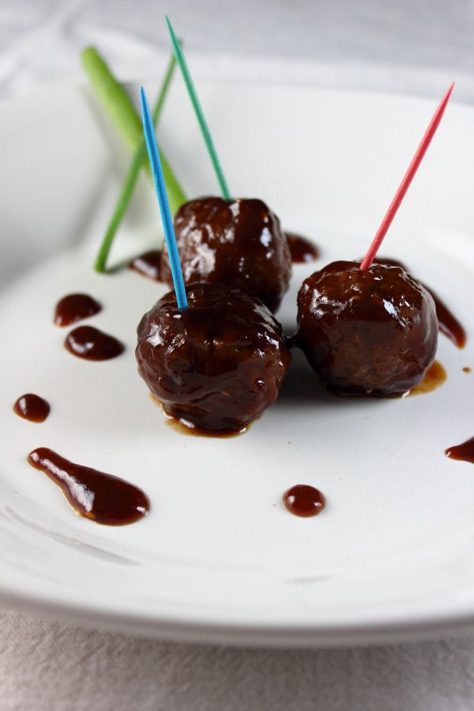 close-up view of three sauce-coated meatballs on a simple white plate. Each meatball is speared with a colored toothpick.