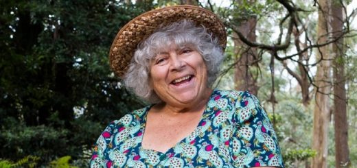 Miriam Margolyes smiles radiantly, sitting in nature in a colourful dress and a straw hat.