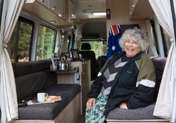 Miriam Margolyes is sitting in a nice caravan, smiling and having tea. There is an Australian flag hanging in the background.