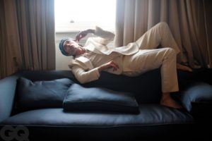 Robert Pattinson lies on top of the backrest of a black sofa by a window. He is wearing a white blazer, slacks, and T-shirt with a blue knitted hat.