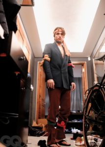 Robert Pattinson is standing in a closet. He is wearing a black blazer and brown pants, with black flip flops. He tied colourful socks around his head, arms, and legs.