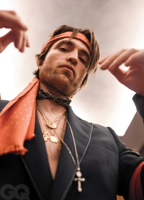 Robert Pattinson is pictured from a lower angle from the chest up. He is wearing a black blazer with no shirt underneath but he has scarves tied around his head and neck and several golden necklaces.