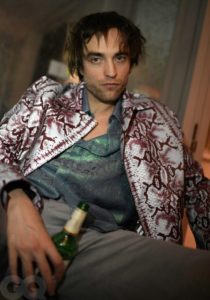 Robert Pattinson is sitting slumped on a chair. He is wearing a snakeskin patterned shirt with a similar glittery t-shirt underneath. He is holding a bottle of beer in his hand.