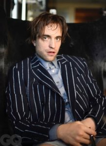 Robert Pattinson is pictured from the waist up sitting on a black couch. He is wearing a black and white striped blazer with a grey button-up and just underwear. His hair could use a comb but he is looking handsome.