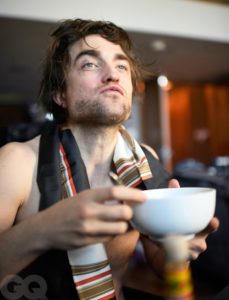 Robert Pattinson is pictured from the waist up wearing no shirt just a silk scarf. He is happily eating cereal out of a plain white bowl.