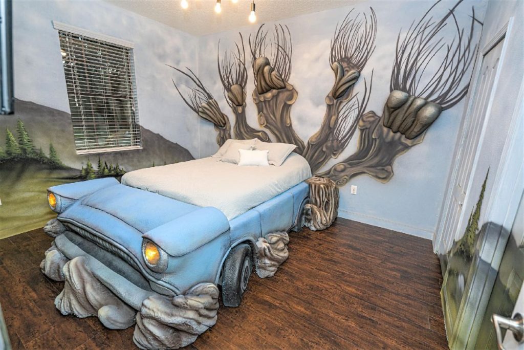 A single bed is pictured in a bedroom. The walls are painted with the landscape of the Hogwarts grounds, with a huge 3-D image of the Whomping Willow at the head of the bed. The bed is custom designed as Arthur Weasley's pale blue Ford Anglia.