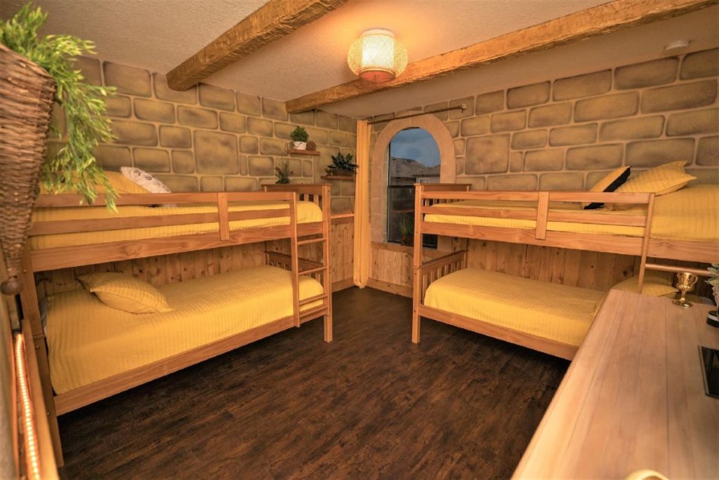 Two sets of bunk beds with yellow bedding are pictured in a cozy Hufflepuff-themed bedroom. There are a few plants and the wallpaper is painted as if it was made of stone.