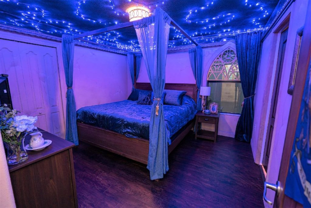 In a bedroom, there is a queen-size four-poster bed with transparent curtains. The ceiling is lit with blue fairy-lights. There are pictures of Rowena Ravenclaw and the Ravenclaw crest on the door.