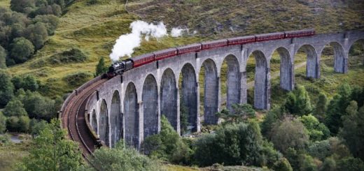 A Hogwarts Express style vintage steam train is travelling down the Glenfinnan Viaduct in the lush, green Highlands.