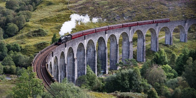 A Hogwarts Express style vintage steam train is travelling down the Glenfinnan Viaduct in the lush, green Highlands.