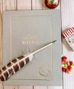 An image of the front cover of the "Harry Potter" Little Keeper Baby Album, which is covered in grey linen with gold embellishments and a quill lying across it.