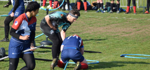 There is one person in a blue jersey on their knees, probably holding a bludger. Another beater is probably trying to take the bludger, while a second beater is running around them. One hoop is standing in the background, and two hoops are on the ground.