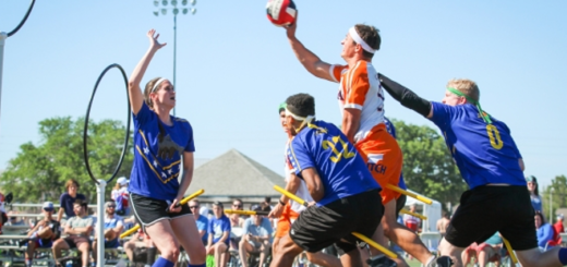 A chaser in an orange jersey is shown attempting to throw the quaffle through the hoops. Three players in blue jerseys are attempting to stop the chaser.