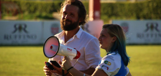 There is a man in white shirt with brown hair and a beard at the left of the image. At the right is a woman in a blue jersey, smaller than the man and holding a megaphone.