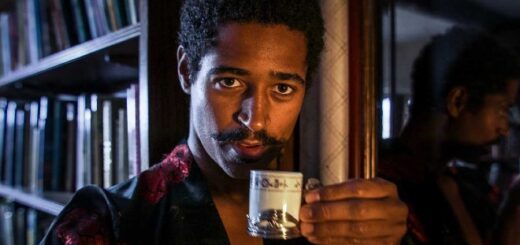 Alfred Enoch is looking directly at the camera while sipping coffee from a fancy cup. He is in character as Lord Henry Wotton and is therefore also sporting a pointy moustache and a luxurious dressing robe.