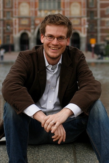 John Green wearing a plaid shirt, brown jacket and blue jeans.