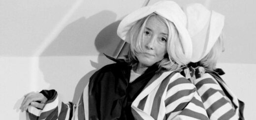 Emma Thompson is pictured in chic stripey clothes and a white hat in a corner of a room. The photo is black and white and she is leaning against a mirror.