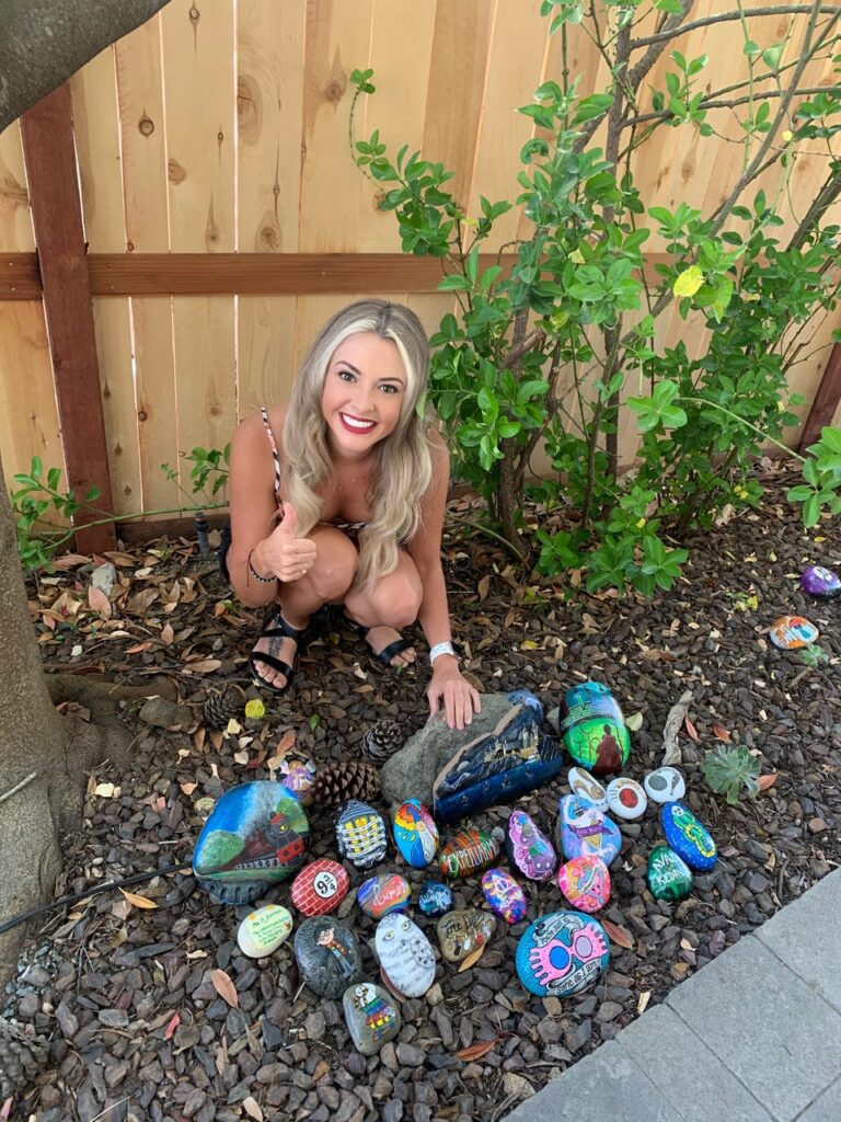 An assortment of painted rocks are arranged on a garden patch. They are painted with Harry Potter thins like Hedwig, Luna's goggles, Hogwarts at night, Harry hovering over a Quidditch pitch on a broomstick, spells, the Platform 9 and 3/4 sign, etc. Next to them,Kristen Newman, the artist is squatting and smiling proudly for the camera.