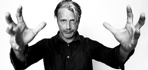 In black and white, Mads Mikkelsen poses in front of a white backdrop, his magnified hands seemngly reaching for the camera.