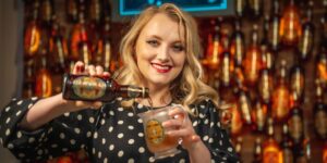 Evanna Lynch is pouring Butterbeer from a bottle into a glass with HP labels with a backdrop of a Butterbeer bar with floating bottles overhead.