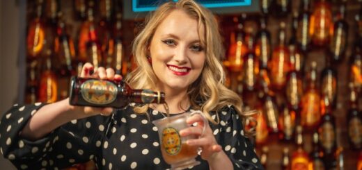 Evanna Lynch is pouring Butterbeer from a bottle into a glass with HP labels with a backdrop of a Butterbeer bar with floating bottles overhead.