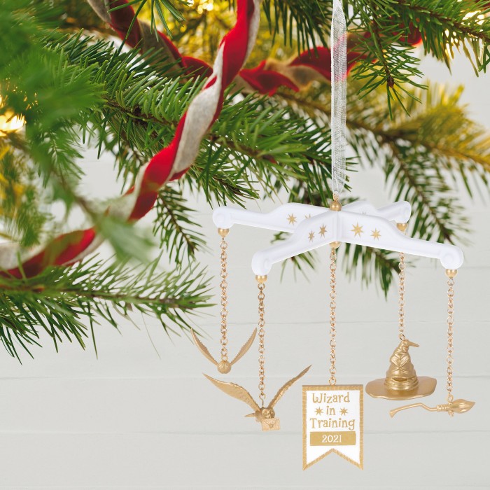 A tiny baby mobile ornament in white and gold is hanging from a Christmas tree branch. A golden snitch, an own, aa sorting hat and a broomstick, as well as a sign saying "Wizard in training 2021" are hanging from the ornament.