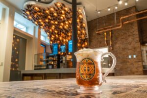 A perfect pour of foamy Butterbeer is placed on a table in the front with a backdrop of a cool bar with Butterbeer bottle lanterns above the counter.