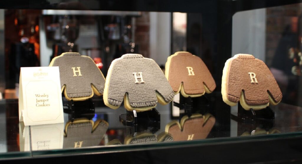 Four adorable sweater-shaped cookies are on display in a snack bar, with initials like the Weasley jumpers in Harry Potter.