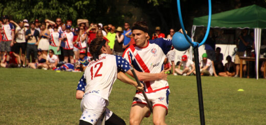 A beater from the United States tries to tackle a beater from Team UK who is holding a blue bludger. There is a hoop next to them.