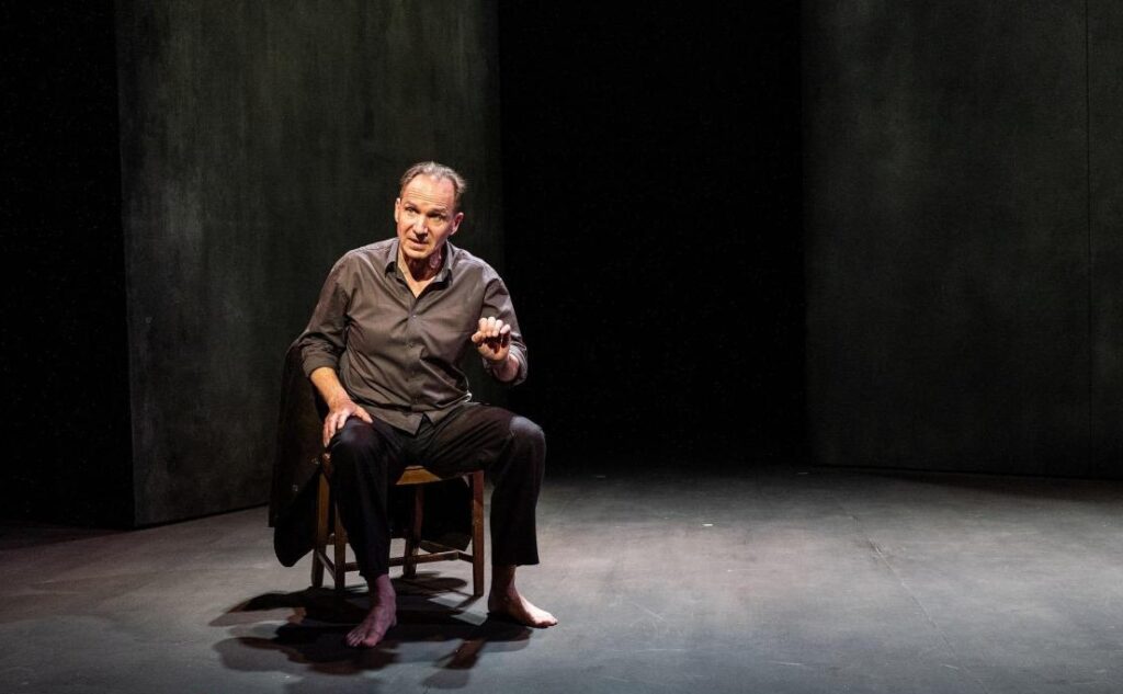 Ralph Fiennes is sitting on a chair barefoot against a dark set, speaking.