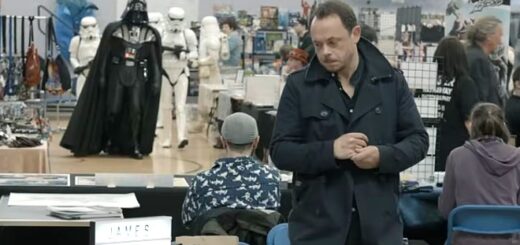 James Payton is standing in a black coat behind a desk at a convention. There is a Darth Vader cosplayer in the background.