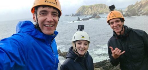 Oliver Phelps, Evanna Lynch, and James Phelps are wearing hard hats and posing in front of the Irish coastline.